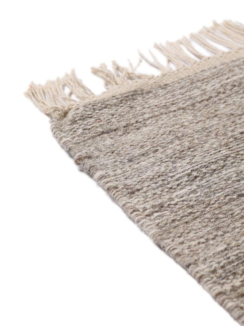 May include: Close-up of a beige woven rug with fringe on the edges. The rug has a subtle texture and a natural, earthy tone.