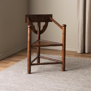 May include: A wooden chair with a triangular seat and a carved back. The chair is made of dark wood and has a rustic look. It is sitting on a gray rug.