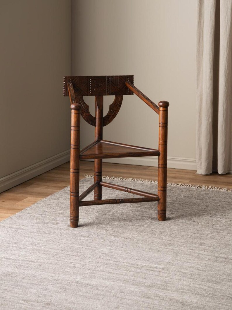 May include: A wooden chair with a triangular seat and a carved back. The chair is made of dark wood and has a rustic look. It is sitting on a gray rug.
