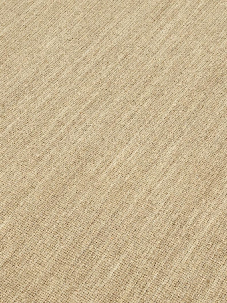 May include: Close-up of a beige woven rug with a subtle diagonal pattern. The texture of the rug is visible, showing the individual strands of the weave.
