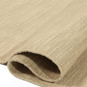 May include: Close-up of a beige rug with a woven texture. The rug is folded in half, showing the edges and the soft, natural fibres.