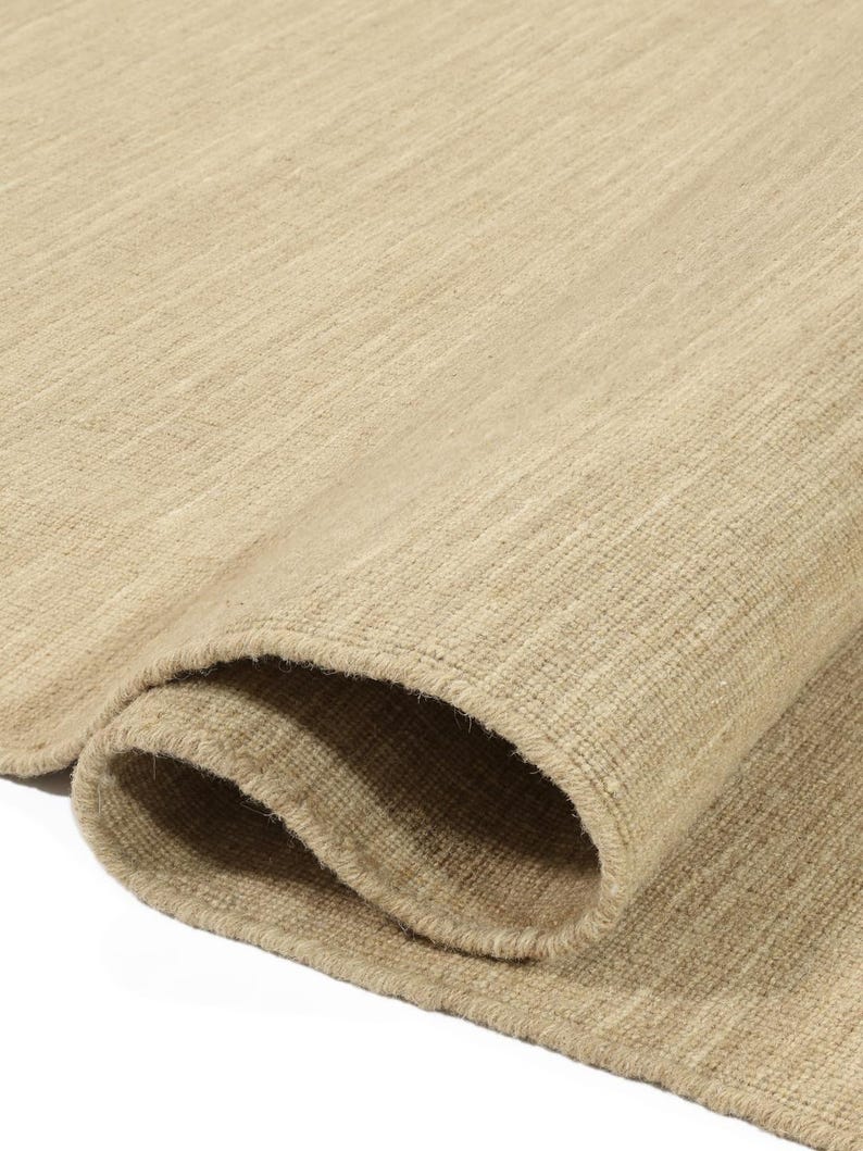 May include: Close-up of a beige rug with a woven texture. The rug is folded in half, showing the edges and the soft, natural fibres.