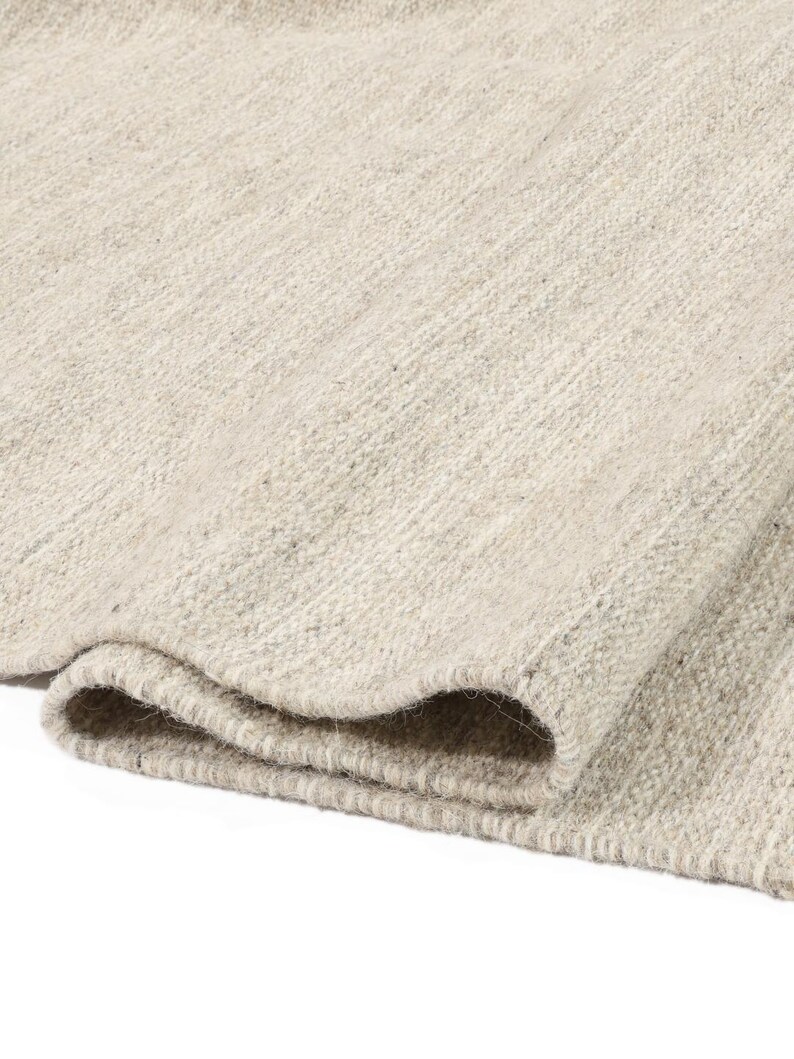 May include: A close-up of a light grey woven rug with a textured surface. The rug is folded in half, showing the edges and the weave.