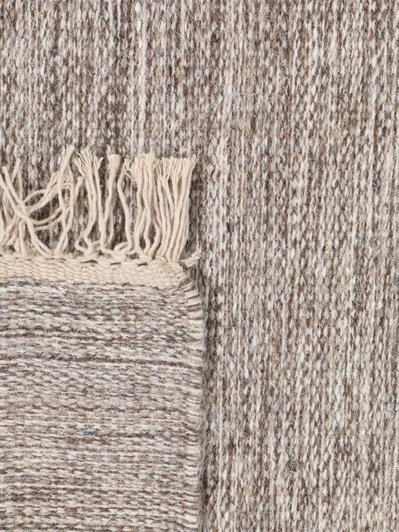 May include: A close-up of a beige and brown woven rug with a fringe border. The rug has a textured, hand-woven look.