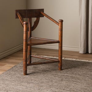 May include: A wooden chair with a carved back and intricate details. The chair has a triangular seat and legs, and is made of dark brown wood. The chair is sitting on a brown rug.