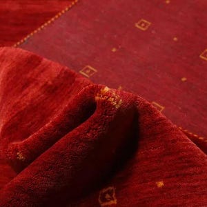 May include: Close-up of a red rug with a subtle pattern of small, gold squares. The rug is folded over, revealing the underside, which is also red.