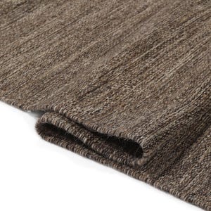 May include: Close-up of a brown and beige woven rug with a textured surface. The rug is folded in half, showing the edge of the fabric.