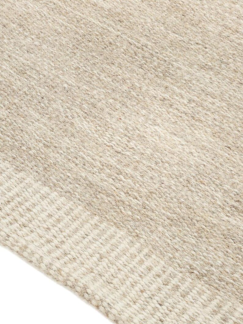 K&ouml;nnte beinhalten: Nahaufnahme eines gewebten Teppichs in Beige und Wei&szlig; mit einer strukturierten Oberfl&auml;che. Der Teppich hat ein dezentes Muster aus Streifen.
