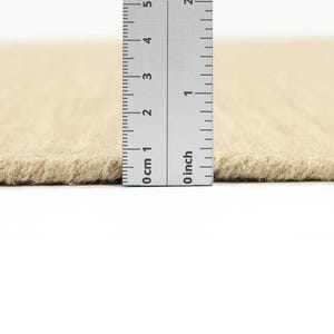 May include: Close-up of a beige rug with a ruler measuring the thickness of the rug. The ruler shows 0 inches and 0 cm.