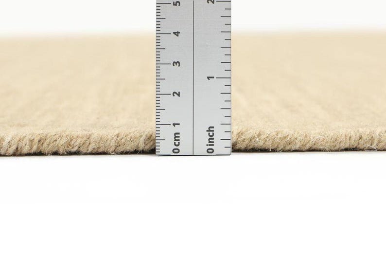 May include: Close-up of a beige rug with a ruler measuring the thickness of the rug. The ruler shows 0 inches and 0 cm.