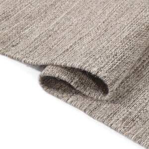May include: Close-up of a light brown woven rug with a textured surface. The rug is folded over on itself, showing the underside of the fabric.