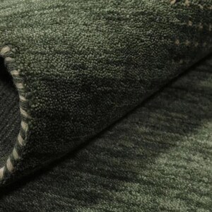 May include: Close-up of a rolled-up, textured green rug. The rug has a dark green color with lighter green and beige accents. The edge of the rug is visible, showing a white stitched border.