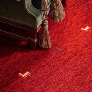 Puede incluir: Una alfombra roja con un patrón texturizado sutil y un contorno de perro marrón claro que se repite en toda la superficie. Una silla de terciopelo verde con borlas marrones se encuentra en la esquina superior derecha de la imagen.