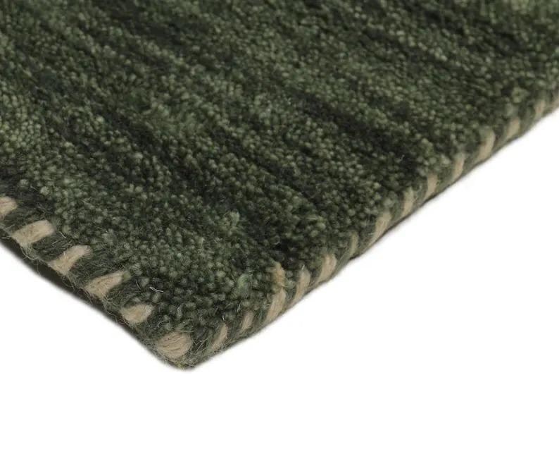 May include: Close-up of a green rug with a textured, ribbed design. The rug has a darker green color with lighter green and beige accents along the edge. The rug's corner is visible against a white background.