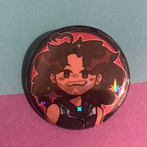 May include: A round, black button with a cartoon illustration of a person with brown hair and a goatee. The illustration has a red border and holographic accents. The background is a mix of blue and pink.