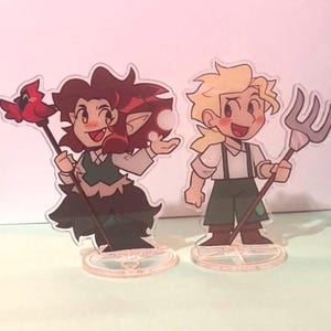 Może przedstawiać: Dwie akrylowe podstawki z postaciami z kreskówek. Jeden ma rude włosy, zieloną kamizelkę i laskę z czerwonym ptakiem. Drugi ma blond włosy, zielone spodnie i widły. Obydwie stoją na przezroczystych podstawach.