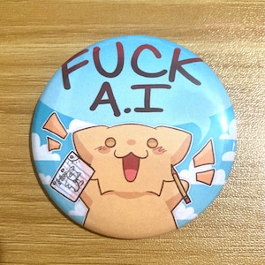 May include: A circular pinback button featuring a light blue sky background with white clouds. The text FUCK A.I is printed in dark brown. A tan cartoon creature holds a pencil and a piece of paper with a drawing of a cat on it.