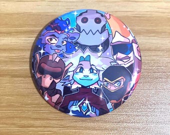 May include: A circular pinback button featuring a colorful, stylized illustration of a group of diverse animated characters. The central figure has light hair and blue skin, surrounded by various figures in helmets and masks, all accented with small white star sparkles.