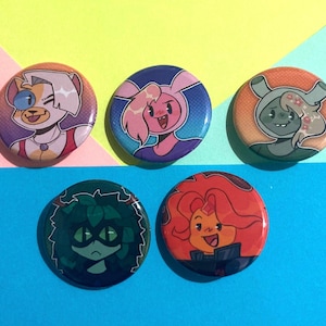 Pode incluir: Cinco botões redondos coloridos com desenhos de personagens de desenhos animados. Os botões apresentam personagens com várias cores, incluindo rosa, laranja, verde e azul. Os botões estão dispostos sobre um fundo amarelo, rosa e azul.