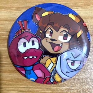 Può includere: Una spilla rotonda con una colorata illustrazione a cartoni animati di tre personaggi su uno sfondo blu scuro. Il design include una creatura rossa, un personaggio dai capelli castani con accenti gialli e una figura robotica azzurra.