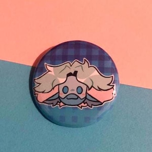 May include: A circular badge featuring a cartoon character with light blue hair and a blue face, set against a blue plaid pattern. The background is split between pink and blue.