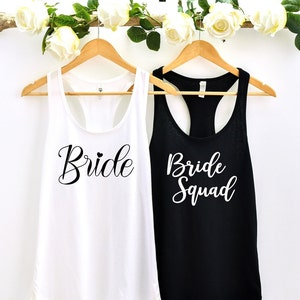 May include: Two tank tops, one white and one black, with the text "Bride" and "Bride Squad" printed on them. The tank tops are hanging on wooden hangers.