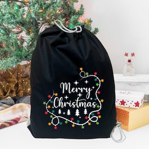 May include: Black drawstring bag with a white "Merry Christmas" design and colorful string lights. The bag is decorated with white stars and small Christmas trees.