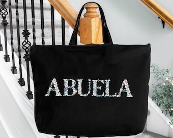 May include: Black canvas tote bag with the word "ABUELA" spelled out in white letters with floral accents.