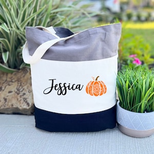 May include: A tri-color grey tote bag with a white panel featuring the name "Jessica" and a stylized orange pumpkin with a white mandala design.