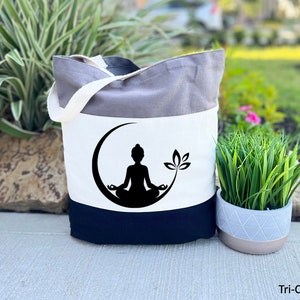 May include: A tri-color tote bag with a grey, white, and black design. The bag features a black silhouette of a person meditating in a crescent moon with a lotus flower.