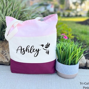 May include: A tri-color tote bag with a pink, white, and burgundy design. The bag features a hummingbird and the name "Ashley" printed in black.