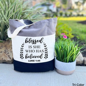 May include: A tri-color gray tote bag with a white panel featuring the text "blessed is she who has believed Luke 1:45".