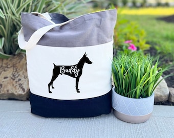 May include: A tri-color grey canvas tote bag with a black silhouette of a Doberman Pinscher and the name "Buddy" printed on the white section of the bag.