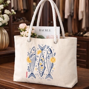 May include: A cream-colored tote bag with white rope handles. The bag features a blue and yellow illustration of three fish with lemon slices. The word "BACRLE" is visible inside the bag, along with flowers.