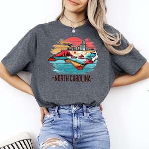 May include: A dark gray t-shirt with a graphic of the state of North Carolina. The graphic is a colorful illustration of the state's outline with a cityscape, mountains, and a sunset in the background. The text "North Carolina" is printed below the graphic.