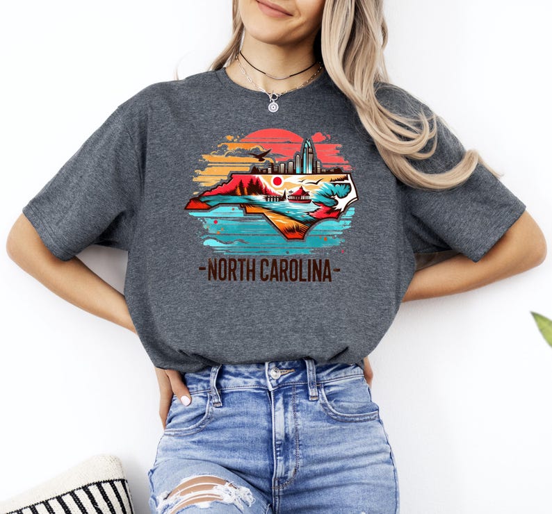 May include: A dark gray t-shirt with a graphic of the state of North Carolina. The graphic is a colorful illustration of the state's outline with a cityscape, mountains, and a sunset in the background. The text "North Carolina" is printed below the graphic.