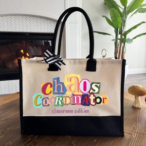 May include: Beige tote bag with black handles and a black base. The bag features the text "Chaos Coordinator" in colorful, cut-out style letters, with "classroom edition" below. A black and white striped bow adorns the top.