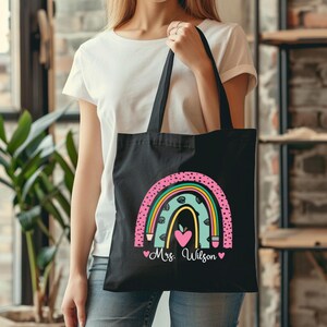 May include: Black tote bag with a rainbow design and the text "Mrs. Wilson" in pink. The rainbow is made up of pink, yellow, green, and blue stripes with black polka dots. There are two pink hearts on either side of the text.