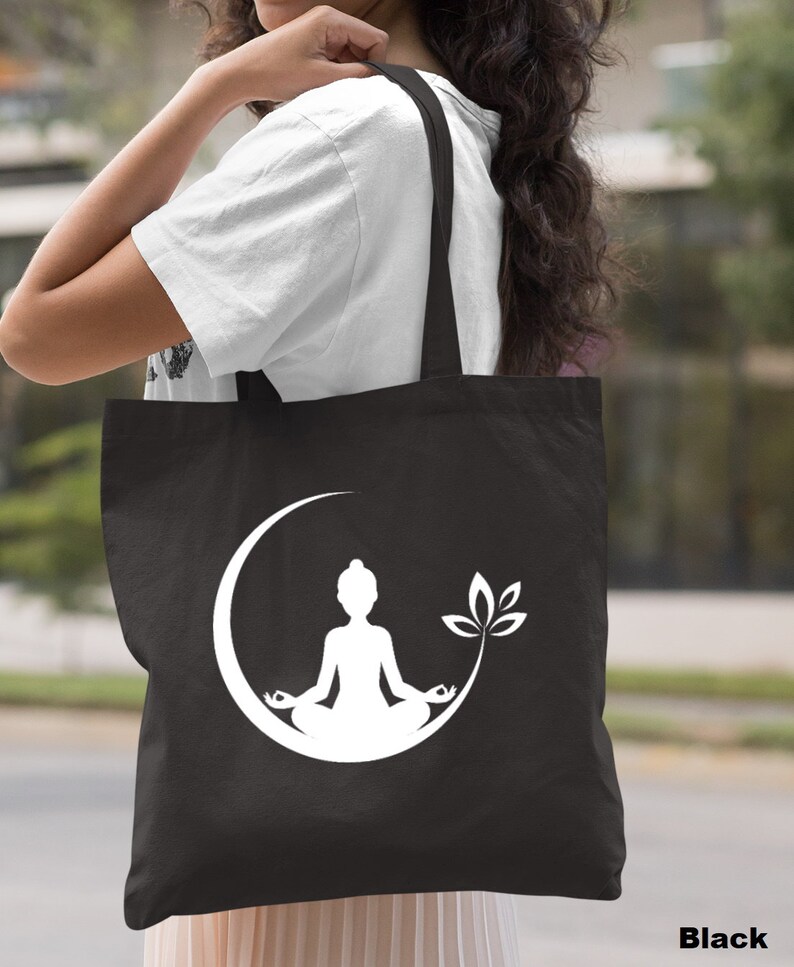 May include: Black canvas tote bag with a white silhouette of a person meditating in a crescent moon with a leaf design.