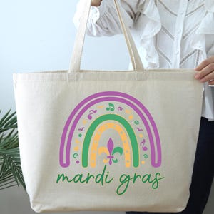 K&ouml;nnte beinhalten: Eine beige Canvas-Tragetasche mit einem Regenbogen-Design in Lila, Gr&uuml;n und Gelb. Der Regenbogen ist mit Noten und Fleur-de-lis-Symbolen verziert. Der Text "Mardi Gras" ist in Gr&uuml;n unter dem Regenbogen geschrieben.