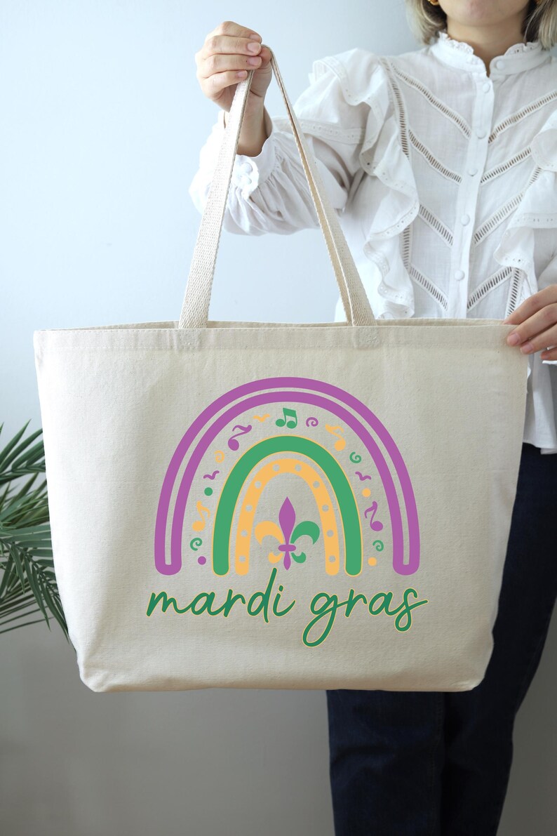 K&ouml;nnte beinhalten: Eine beige Canvas-Tragetasche mit einem Regenbogen-Design in Lila, Gr&uuml;n und Gelb. Der Regenbogen ist mit Noten und Fleur-de-lis-Symbolen verziert. Der Text "Mardi Gras" ist in Gr&uuml;n unter dem Regenbogen geschrieben.