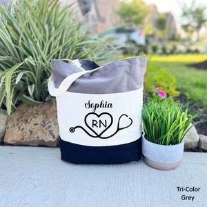 May include: A white canvas tote bag with a gray and black stripe design. The bag has a stethoscope and heart with the letters "RN" inside. The name "Sophia" is printed above the heart.