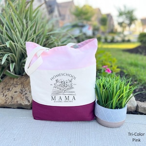 Peut inclure: Un sac fourre-tout rose tricolore avec le texte "Homeschool Mama Raising Arrows" imprim&eacute; sur le devant. Le sac a un dessus rose, un centre blanc et un fond rose fonc&eacute;. Le sac est en toile et poss&egrave;de des poign&eacute;es blanches.