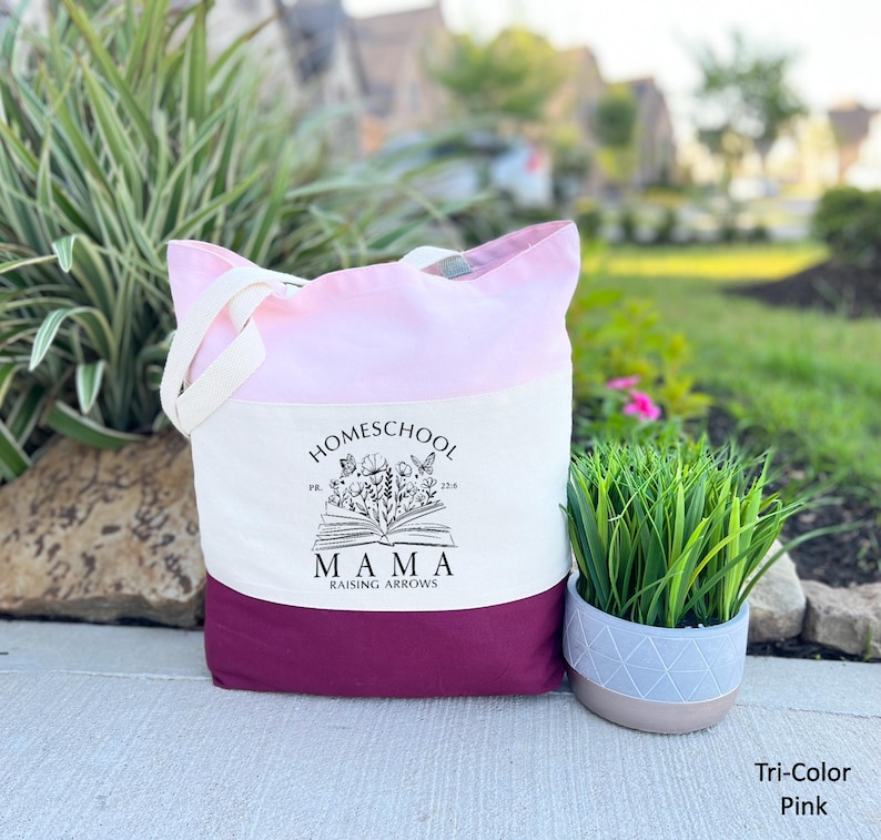 Peut inclure: Un sac fourre-tout rose tricolore avec le texte "Homeschool Mama Raising Arrows" imprim&eacute; sur le devant. Le sac a un dessus rose, un centre blanc et un fond rose fonc&eacute;. Le sac est en toile et poss&egrave;de des poign&eacute;es blanches.