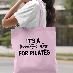 May include: Light pink tote bag with the text "IT'S A beautiful day FOR PILATES" in black. The bag has long straps and is made of a soft material. The words "Light Pink" are in the bottom right corner.