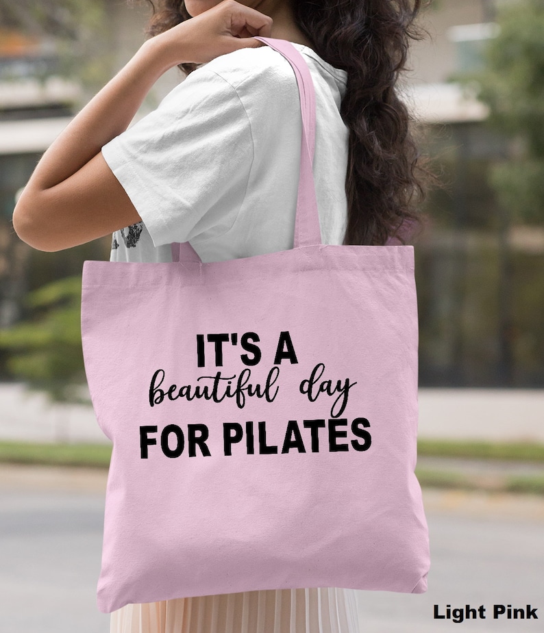 May include: Light pink tote bag with the text "IT'S A beautiful day FOR PILATES" in black. The bag has long straps and is made of a soft material. The words "Light Pink" are in the bottom right corner.
