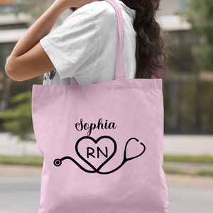 May include: Light pink tote bag with a black stethoscope and heart design. The heart has the letters "RN" inside. The text "Sophia" is printed above the heart.