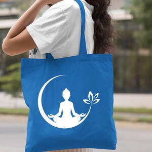 May include: A royal blue tote bag with a white silhouette of a person meditating in a crescent moon with a lotus flower. The bag has two white straps.