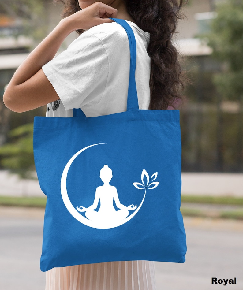May include: A royal blue tote bag with a white silhouette of a person meditating in a crescent moon with a lotus flower. The bag has two white straps.