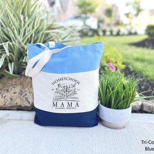 Peut inclure: Un sac fourre-tout bleu tricolore avec des poign&eacute;es blanches. Le sac pr&eacute;sente un haut bleu clair, un centre blanc avec un motif floral et un livre, et un bas bleu marine. Le texte "Homeschool Mama Raising Arrows" est imprim&eacute; sur le devant.